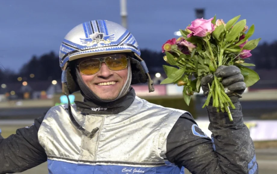 Carl Johan Jepson i silverfärgad kuskdress och hjälm ler och håller upp en segerbukett på travbanan