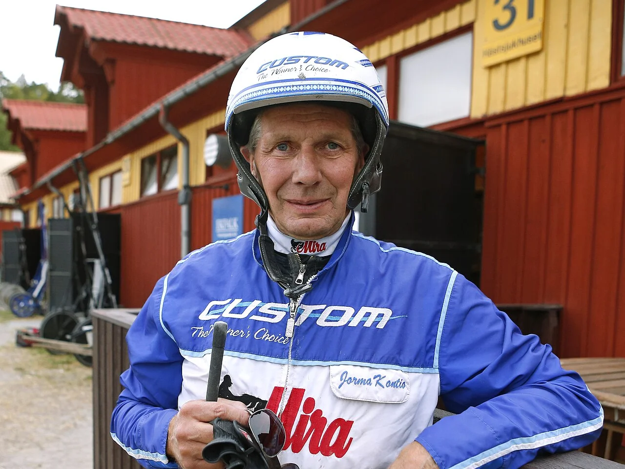 Jorma Kontio i blå och vit kuskdress och hjälm framför ett rött stallhus